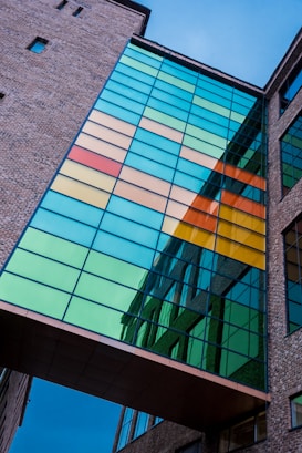 A modern building exterior featuring a glass bridge with vibrant colored panels in shades of blue, green, yellow, orange, and red. The structure is framed by brickwork, and the panels reflect the surrounding architecture and sky.