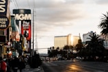 A bustling city street with various people walking on the sidewalk. Prominent buildings and billboards are visible, including signs for MGM, Coca-Cola, and Brad Garrett’s Comedy Club. The image captures the golden reflection of sunlight on the Mandalay Bay hotel, adding a warm glow to the scene. Palm trees line the street, and scattered clouds fill the sky.