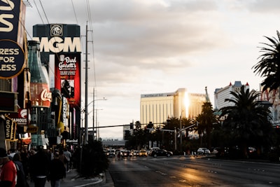 A bustling city street with various people walking on the sidewalk. Prominent buildings and billboards are visible, including signs for MGM, Coca-Cola, and Brad Garrett’s Comedy Club. The image captures the golden reflection of sunlight on the Mandalay Bay hotel, adding a warm glow to the scene. Palm trees line the street, and scattered clouds fill the sky.