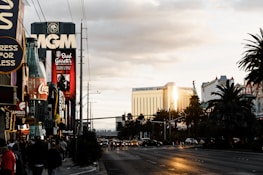 A bustling city street with various people walking on the sidewalk. Prominent buildings and billboards are visible, including signs for MGM, Coca-Cola, and Brad Garrett’s Comedy Club. The image captures the golden reflection of sunlight on the Mandalay Bay hotel, adding a warm glow to the scene. Palm trees line the street, and scattered clouds fill the sky.