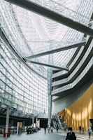 An expansive interior with a high glass ceiling and curved, modern architectural lines. The space features a large atrium with a combination of metal beams and glass, creating a bright and airy environment. People are walking and sitting in the open space, and an escalator is visible on one side.