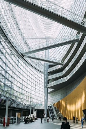 An expansive interior with a high glass ceiling and curved, modern architectural lines. The space features a large atrium with a combination of metal beams and glass, creating a bright and airy environment. People are walking and sitting in the open space, and an escalator is visible on one side.