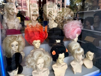 Mannequin heads with various styles of wigs are displayed in a store window. The wigs come in multiple colors, including blonde, black, pink, and bright red. The wigs are styled in elaborate and voluminous fashion, giving a vibrant and eclectic appearance. The reflection of buildings and trees outside can be seen on the window glass.