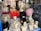 Mannequin heads with various styles of wigs are displayed in a store window. The wigs come in multiple colors, including blonde, black, pink, and bright red. The wigs are styled in elaborate and voluminous fashion, giving a vibrant and eclectic appearance. The reflection of buildings and trees outside can be seen on the window glass.