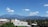 A scenic view featuring a range of blue mountains under a bright, clear sky with fluffy white clouds. Below the mountains, there are several buildings with metal rooftops surrounded by lush green foliage. Palm trees are also visible, adding to the tropical atmosphere.