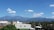 A scenic view featuring a range of blue mountains under a bright, clear sky with fluffy white clouds. Below the mountains, there are several buildings with metal rooftops surrounded by lush green foliage. Palm trees are also visible, adding to the tropical atmosphere.