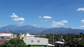 A scenic view featuring a range of blue mountains under a bright, clear sky with fluffy white clouds. Below the mountains, there are several buildings with metal rooftops surrounded by lush green foliage. Palm trees are also visible, adding to the tropical atmosphere.