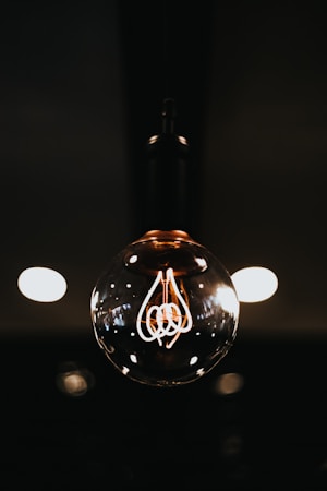 A glowing Edison-style light bulb hangs against a dark background. The filament inside the bulb is clearly visible and emits a warm, inviting glow. The light reflects gently off the glass surface of the bulb, and out-of-focus light spots in the background add depth to the composition.