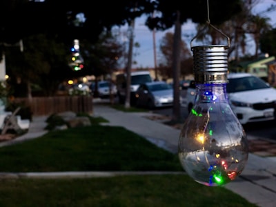 A decorative light bulb hangs outdoors, reflecting colorful lights within its glass casing. Behind it, a suburban street scene unfolds with parked cars and a lush green lawn. The sky is overcast, with the light of early evening creating a cozy, tranquil atmosphere.