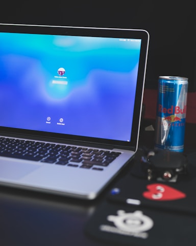 A laptop is displaying a stylish lock screen with a blurred blue background. To the right, a can of Red Bull is visible on the surface next to some electronic accessories, including a computer mouse resting on a black mousepad with a red heart emblem. The environment suggests a workspace or study area.