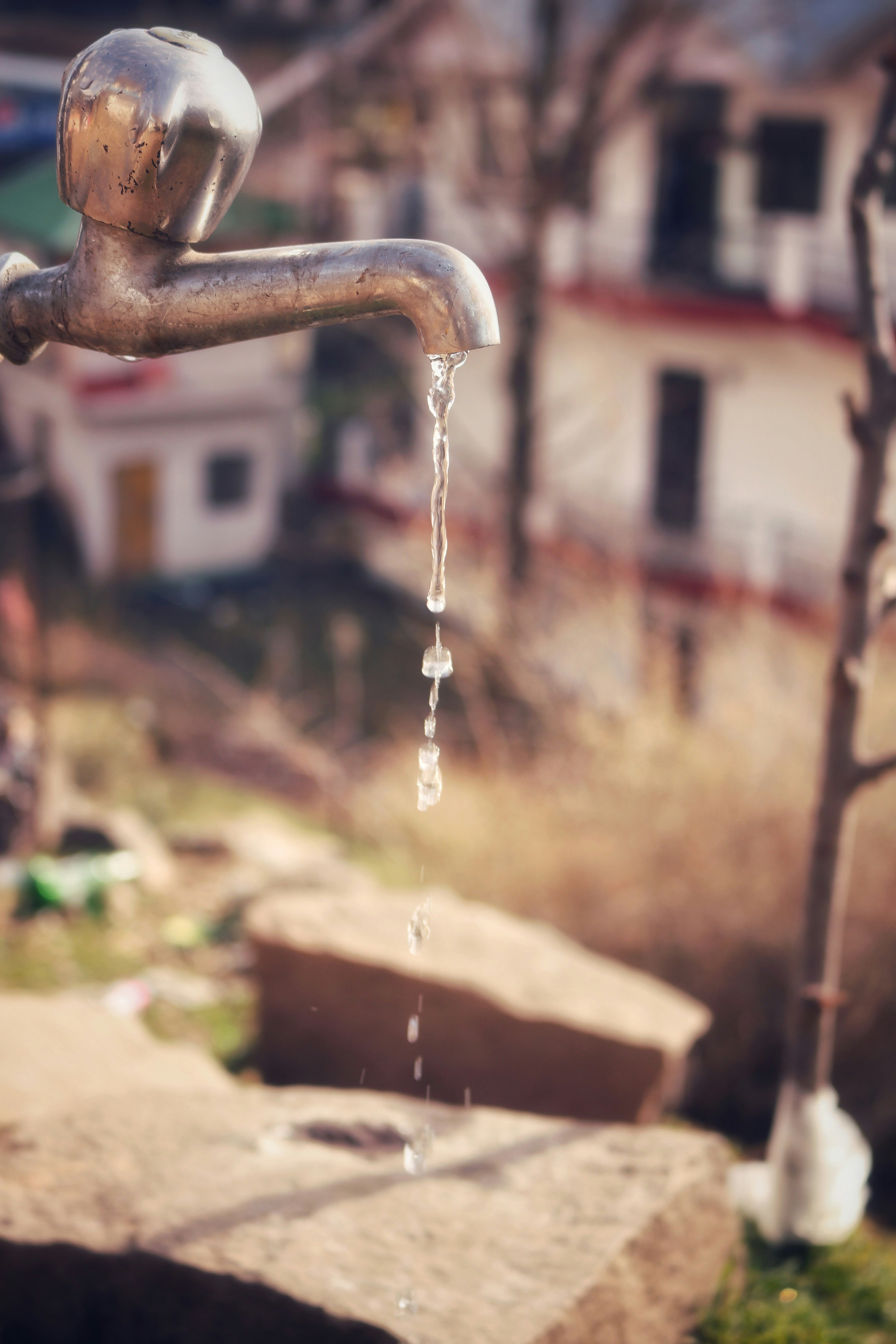 A close-up of a metal faucet with water trickling down from its spout. The background appears to be outdoors with blurred structures and greenery, creating a soft focus effect.