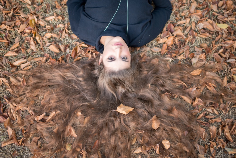 Flay lay photographie d’une femme brune aux cheveux longs portant un haut à col rond noir allongé sur un champ avec des feuilles sèches