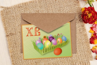 A greeting card with decorative eggs placed on a background of green grass and an envelope partially visible. The background features a textured fabric, and various colorful flowers are scattered on the right side.