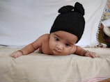 A baby lying on their stomach, wearing a black knitted hat with two round protrusions. The baby is on a beige blanket with a white background, and has beaded bracelets on both wrists.