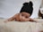 A baby lying on their stomach, wearing a black knitted hat with two round protrusions. The baby is on a beige blanket with a white background, and has beaded bracelets on both wrists.