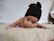 A baby lying on their stomach, wearing a black knitted hat with two round protrusions. The baby is on a beige blanket with a white background, and has beaded bracelets on both wrists.
