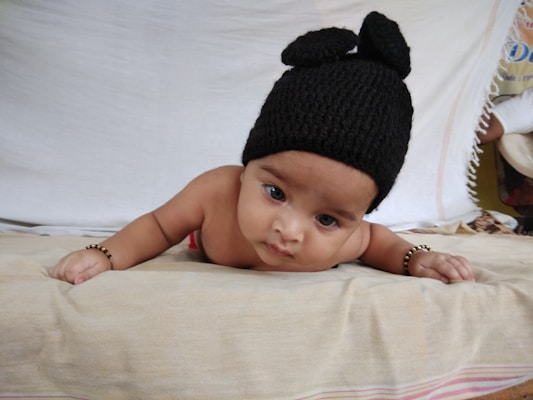 A baby lying on their stomach, wearing a black knitted hat with two round protrusions. The baby is on a beige blanket with a white background, and has beaded bracelets on both wrists.
