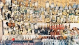 A vast collection of traditional handicrafts and artifacts is displayed. This includes intricately crafted masks, bells, carved statues, and various small figurines, all arranged neatly on shelves and hanging from above. The items appear to be made from materials like wood, metal, and clay, showcasing a mix of cultural and artistic designs.