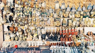 A vast collection of traditional handicrafts and artifacts is displayed. This includes intricately crafted masks, bells, carved statues, and various small figurines, all arranged neatly on shelves and hanging from above. The items appear to be made from materials like wood, metal, and clay, showcasing a mix of cultural and artistic designs.