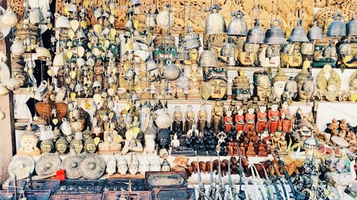 A vast collection of traditional handicrafts and artifacts is displayed. This includes intricately crafted masks, bells, carved statues, and various small figurines, all arranged neatly on shelves and hanging from above. The items appear to be made from materials like wood, metal, and clay, showcasing a mix of cultural and artistic designs.
