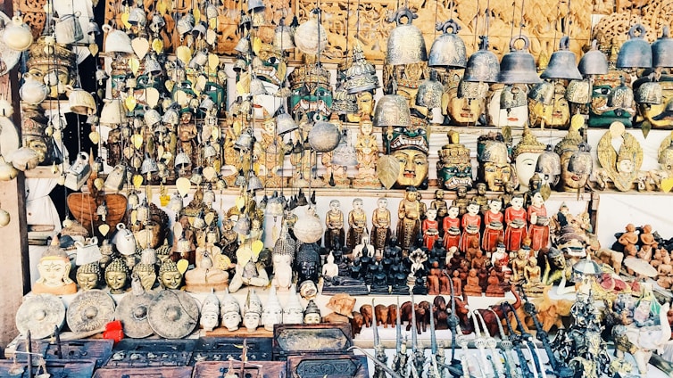 A vast collection of traditional handicrafts and artifacts is displayed. This includes intricately crafted masks, bells, carved statues, and various small figurines, all arranged neatly on shelves and hanging from above. The items appear to be made from materials like wood, metal, and clay, showcasing a mix of cultural and artistic designs.