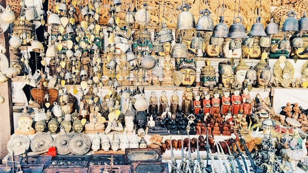 A vast collection of traditional handicrafts and artifacts is displayed. This includes intricately crafted masks, bells, carved statues, and various small figurines, all arranged neatly on shelves and hanging from above. The items appear to be made from materials like wood, metal, and clay, showcasing a mix of cultural and artistic designs.