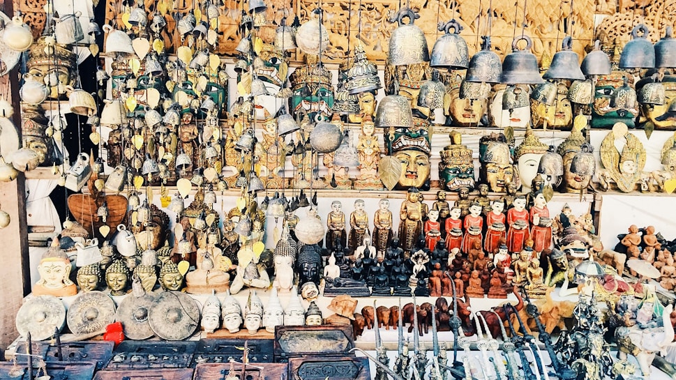 A vast collection of traditional handicrafts and artifacts is displayed. This includes intricately crafted masks, bells, carved statues, and various small figurines, all arranged neatly on shelves and hanging from above. The items appear to be made from materials like wood, metal, and clay, showcasing a mix of cultural and artistic designs.