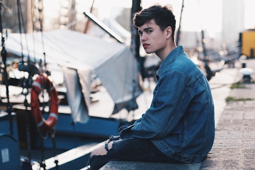 A young person sits on a concrete ledge near a harbor, wearing a denim jacket and ripped jeans. The background features sailboats and maritime equipment, including a life ring.