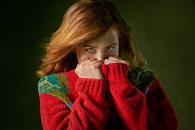 Uma jovem mulher ruiva e de casaco vermelho com as mãos cobrindo o rosto expressando vergonha