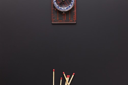 A compass is positioned at the top against a dark background, with several wooden matches arranged at the bottom, their red tips pointing upwards.