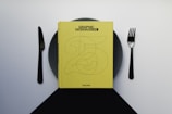 A yellow book titled 'Graphic Design Now' is placed on a dark plate, with a knife on the left and a fork on the right, resembling a dining setup. The background is a gradient of light to dark gray.
