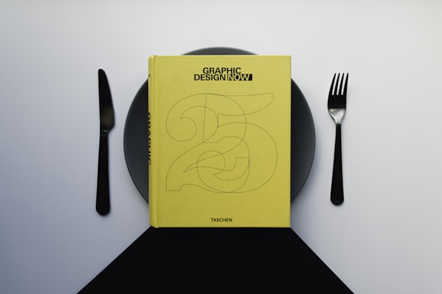 A yellow book titled 'Graphic Design Now' is placed on a dark plate, with a knife on the left and a fork on the right, resembling a dining setup. The background is a gradient of light to dark gray.