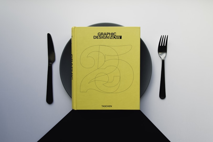 A yellow book titled 'Graphic Design Now' is placed on a dark plate, with a knife on the left and a fork on the right, resembling a dining setup. The background is a gradient of light to dark gray.