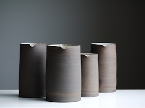 Four tall, cylindrical ceramic pitchers with a minimalist design are arranged on a white surface against a muted gray background. Each pitcher has a matte finish with slight variations in tone, ranging from dark to light grayish-brown.