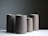 Four tall, cylindrical ceramic pitchers with a minimalist design are arranged on a white surface against a muted gray background. Each pitcher has a matte finish with slight variations in tone, ranging from dark to light grayish-brown.
