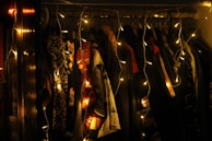 A closet full of various hanging clothes illuminated by warm-colored fairy lights. The lights create a cozy and inviting atmosphere, with garments of different colors and patterns visible in the dim lighting.