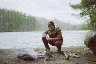 A person is crouching by a small campfire near a lake, surrounded by a forest. The individual is wearing a brown jacket and appears focused on maintaining the fire. The setting is serene, with the lake calm and trees in the background.