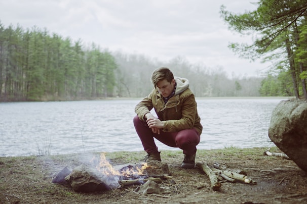 A person is crouching by a small campfire near a lake, surrounded by a forest. The individual is wearing a brown jacket and appears focused on maintaining the fire. The setting is serene, with the lake calm and trees in the background.