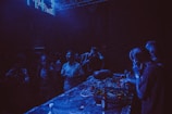 A dimly lit nightclub with people gathered around a DJ booth. The environment is filled with blue and purple lighting. The DJ is wearing headphones and operating the equipment, while attendees are enjoying the music and socializing nearby.