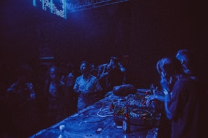 A dimly lit nightclub with people gathered around a DJ booth. The environment is filled with blue and purple lighting. The DJ is wearing headphones and operating the equipment, while attendees are enjoying the music and socializing nearby.