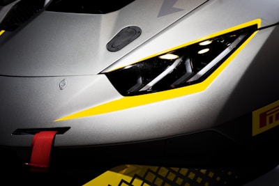 A close-up of the front part of a gray sports car with sleek, aerodynamic lines. The car features a prominent headlight design, accented with yellow details around the edges. A red tow hook is visible on the bumper, suggesting a racing or high-performance variant. The overall design conveys a sense of speed and precision.