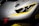 A close-up of the front part of a gray sports car with sleek, aerodynamic lines. The car features a prominent headlight design, accented with yellow details around the edges. A red tow hook is visible on the bumper, suggesting a racing or high-performance variant. The overall design conveys a sense of speed and precision.