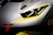 A close-up of the front part of a gray sports car with sleek, aerodynamic lines. The car features a prominent headlight design, accented with yellow details around the edges. A red tow hook is visible on the bumper, suggesting a racing or high-performance variant. The overall design conveys a sense of speed and precision.