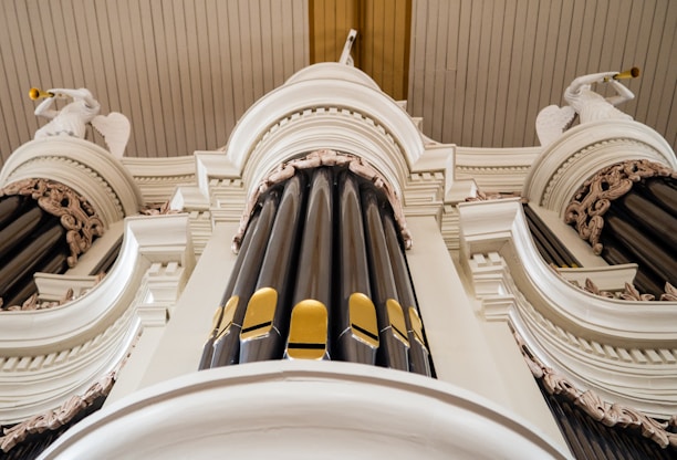 An ornate pipe organ with intricate detailing is depicted. The image captures the impressive vertical structure of the organ pipes, surrounded by decorative carvings and embellishments. Two figures, possibly angels, emerge from the sides, each holding a trumpet.