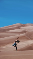 woman standing on right foot and raising hands at the bottom of sand dunes during daytime