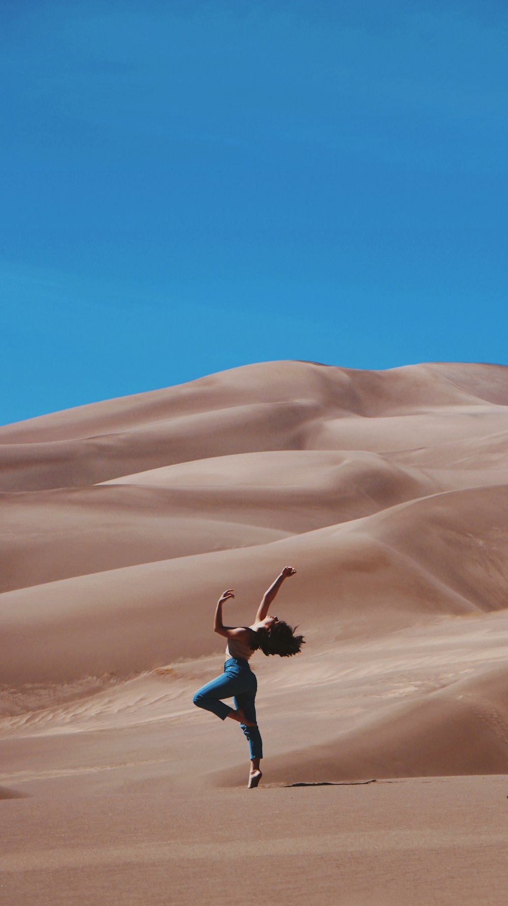 woman standing on right foot and raising hands at the bottom of sand dunes during daytime
