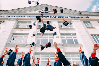A group of graduates in blue gowns with red accents throw their caps into the air in front of a large building. The building has classical architectural features, such as columns and large windows, with some signage in a foreign script at the top.