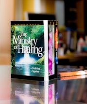 A set of colorful books is neatly arranged on a reflective surface, with the most prominent book titled 'The Ministry of Healing'. The background features a blurred indoor setting, likely a room or office.