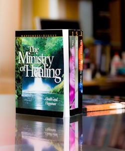 A set of colorful books is neatly arranged on a reflective surface, with the most prominent book titled 'The Ministry of Healing'. The background features a blurred indoor setting, likely a room or office.