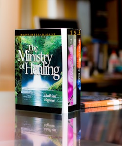 A set of colorful books is neatly arranged on a reflective surface, with the most prominent book titled 'The Ministry of Healing'. The background features a blurred indoor setting, likely a room or office.